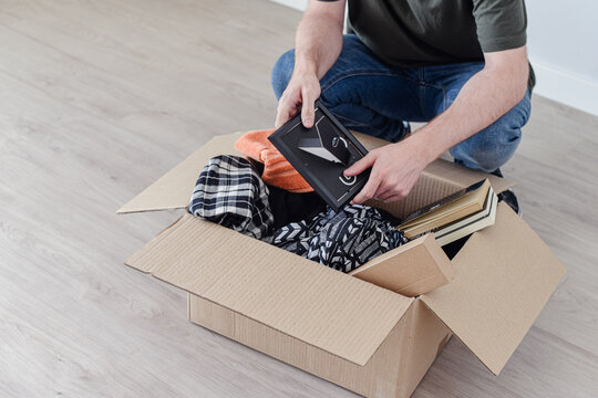 Un Hombre Joven Agachado Buscando Cosas En Una Caja De Mudanza, Llena De Ropa, Libros Y Un Marco De Foto.