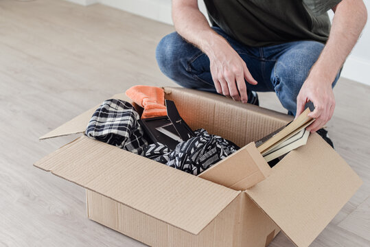 Un hombre joven agachado buscando cosas en una caja de mudanza, llena de ropa, libros y un marco de foto.