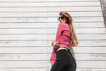 Fashionable beautiful slender girl model with trendy sunglasses in fashion bright summer clothes with a pink top and a black skirt poses near a white wooden wall