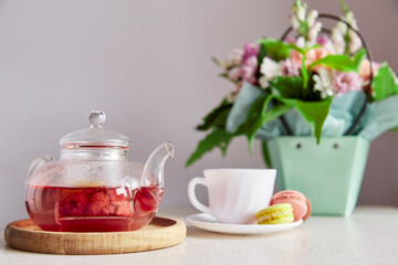 Cozy tea time - cup of tea with macarons, glass teapot, candle and bouquet of flowers. Herbal raspberry natural tea. Aesthetic home with tea and dessert. Self care, relaxing, wellness lifestyle.