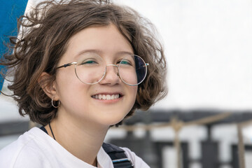 Portrait of smiling teenage girl with curly hair wearing fashionable glasses in city park on sunny summer day, children's or teen fashion, sense of freedom