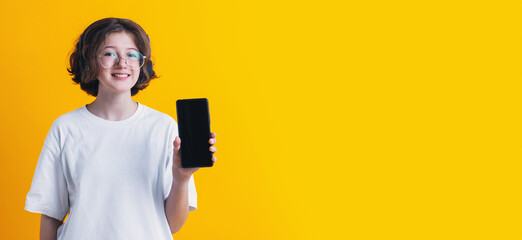 girl in glasses and headphones shows in her hand smartphone with empty dark screen on colored background, communication, design of advertising banners, business cards and mockup, copy space