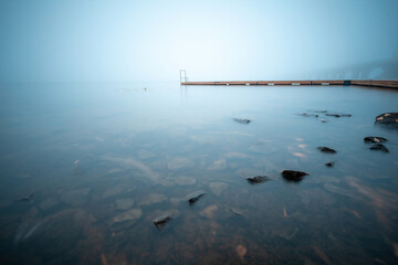 Wooden jetty on a foggy river with stones in the water on a foreground. Minimalist long exposure landscape.