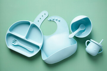 Silicone baby feeding set. Flat lay silicone bib, plate, cutlery and cup on green background. Baby...