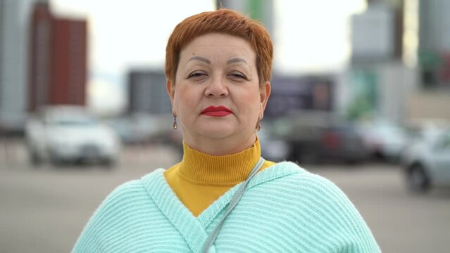 Portrait of a middle-aged woman with a short haircut and brightly painted lips looking at the camera against the backdrop of cars and houses