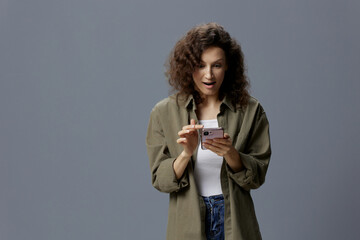 Shocked excited confused curly beautiful woman in casual khaki green shirt get shock info from message using phone posing isolated on gray blue background. Social Media Influencer concept. Copy space
