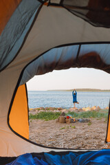 view from a tent on the shore of a lake or sea, a woman collects firewood or does yoga and...