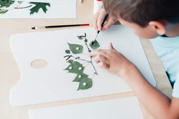 Little child sitting by desk and making picture from dry birch leaves