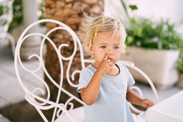 Little girl picks her nose while sitting on a wrought-iron chair at the table. High quality photo