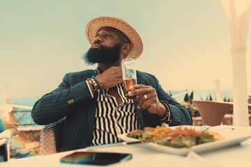 A bearded black man in a casual attire and a straw hat is adjusting his shirt lapel while drinking...