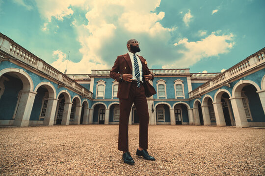 A stately mature bald bearded black man in a fashionable tailored dark-red suit and eyeglasses is standing in the center of a courtyard of a blue antique building with a teal sky; a wide-angle view