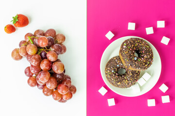 Healthy and unhealthy food, flat layer, two-color background, chocolate donuts with sprinkles on a fuchsia background, juicy delicious grapes, strawberries on white, natural sugar vs harmful processed