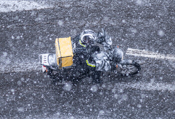 food delivery courier rides a black motorcycle under the snowfall with a yellow backpack. View from above
