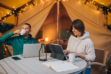 Happy family couple freelancers working laptop on a cozy glamping tent in summer evening. Luxury camping tent for outdoor holiday and vacation. Lifestyle concept