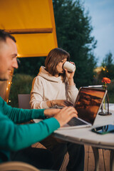 Woman freelancer drink coffee and working laptop near colleague on  cozy glamping tent in summer evening. Luxury camping tent for outdoor holiday and vacation. Lifestyle concept