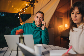 Woman freelancer working laptop and making notes while colleague talking phone sitting in cozy glamping tent in summer evening. Luxury camping tent for outdoor holiday and vacation. Lifestyle concept