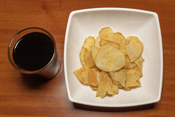 Overhead shot of a white plate with chips next to a glass with black soda on a wooden table