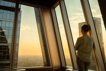 Back view of woman looking at cityscape through window of skyscraper. Summer time, sunset, warm...