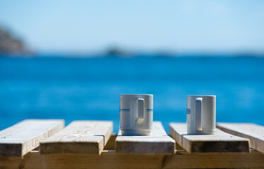 Two mugs on a wooden table by the sea.