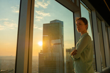 Portrait of smiling woman looking at cityscape through window of skyscraper. Summer time, sunset, lens flare. Relax, leisure time, success, opportunity, sightseeing, discover and future concept