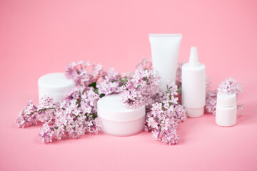 Obraz na płótnie Canvas Face and body skin care. A set of cosmetic creams and balms in white tubes and cans on a coral background with sprigs of lilac flowers. Spa treatments for home care. Home rejuvenation and moisturizing