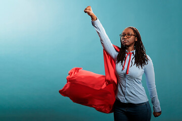 Mighty and brave african american superhero woman posing as justice defender while acting like flying on blue background. Young adult strong person wearing red hero cloak. Studio shot. © DC Studio