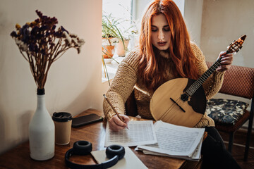 Redhead woman playing acoustic Domra Folk Musical Instrument and looking into note. Young woman taking music lessons at home