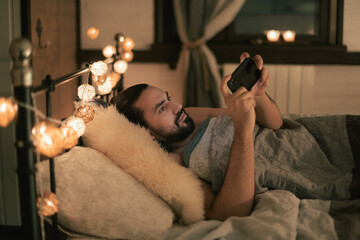 Young man with phone in bed in the evening. Evening leisure at home