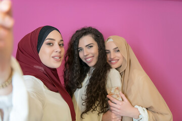 group of beautiful muslim women two of them in fashionable dress with hijab using mobile phone while taking selfie picture isolated on pink background representing modern islam fashion technology 