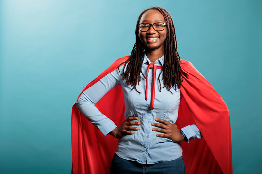 Proud superhero woman with superpower abilities wearing mighty hero cloak while smiling confident at camera. Joyful happy smiling young adult person posing as justice defender on blue background.