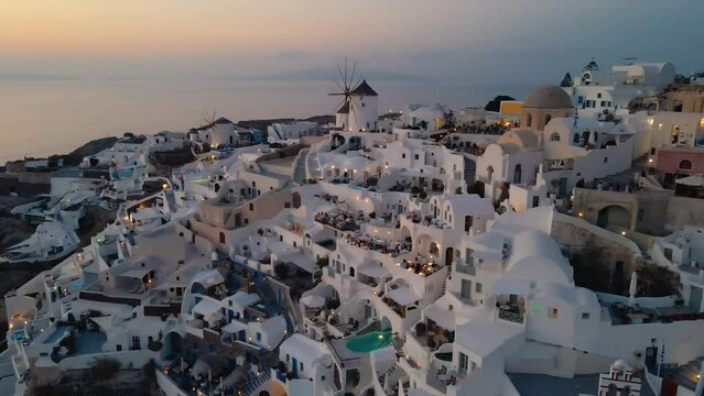Cinematic Aerial Shot of Oia Village, Santorini, Greece - Cyclades - close-up on famous Windmills silhouetting against the Aegean Sea, while tourists enjoy sunset from typical whitewashed houses
