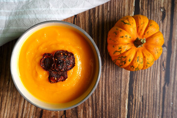 pumpkin soup puree in a round plate with slices of dried tomato and a small pumpkin stand next to each other on a brown wooden table. view from above. vegan