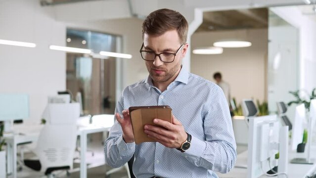 Close up portrait of young Caucasian man standing in cabinet in modern company looking away while browsing on tablet. Handsome male manager using device at workplace. Office worker Businessman concept