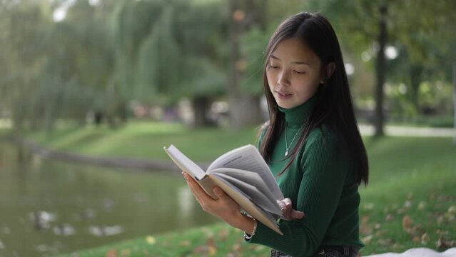 Young smiling intelligent woman turning pages reading book in summer park at pond. Portrait of confident relaxed happy Asian lady enjoying hobby outdoors on sunny day. Lifestyle concept