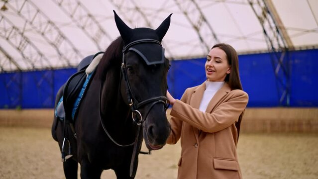 Beautiful brunette woman in beige jacket holding a beautiful black horse. Lady leading a graceful animal by the drill-hall. Blurred backdrop.