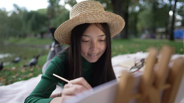 Inspired talented Asian young woman painting on canvas lying on summer meadow in park smiling. Portrait of happy cheerful artist enjoying hobby outdoors creating masterpiece