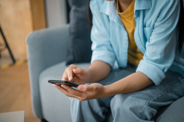 Fototapeta premium Smiling young asian woman using mobile phone checking social media,app playing game, shopping online, ordering delivery relax while sitting on a couch at home with laptop computer