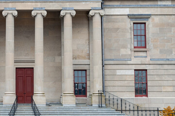 The exterior facade of a historic government building with large vintage marble pillars or columns, a red door, black rails, and a brick entrance leading up steps to the large antique stone building.
