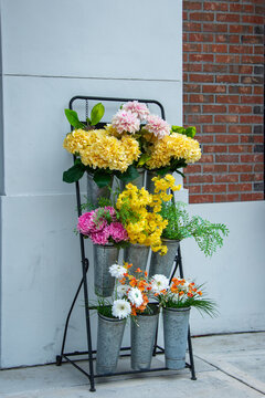 A Sidewalk Stand With Multiple Containers Of Colorful Cut Flower Bouquets On Display For Sale. There Are Yellow, Pink, White Orange And Green Blooms. The Metal Container Has Aluminum Vases For Storage