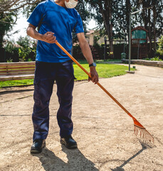 Young latino janitorial worker in blue uniform sweeping with orange rake in the park 