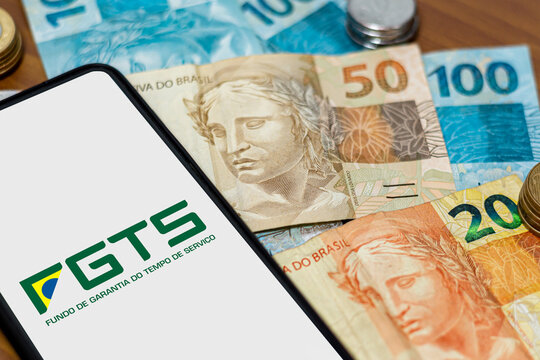 Sao Paulo, Brazil. SEPTEMBER 19, 2022: FGTS logo on smartphone screen with multiple coins and moneys around. FGTS is the Brazilian worker's guarantee fund. Payment by the Brazilian government