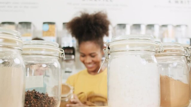 Close-up and selective focus on reusable glass jars, two young female customers choose and shop for organic products in refill stores, zero-waste grocery, and plastic-free, eco-friendly lifestyles.