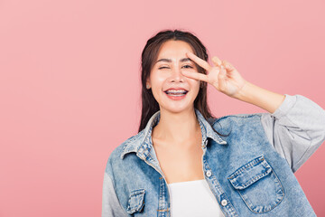 Asian happy portrait beautiful cute young woman wear denim smile standing showing finger making v-sign victory symbol near eye looking to camera studio shot isolated on pink background with copy space