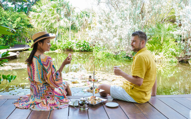 Afternoon tea at a water pond, high tea in a tropical garden in Thailand. Couple of men and women...