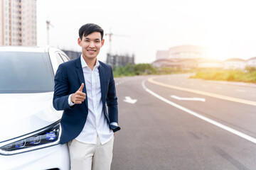 Asian man looks on camera while standing near new electric car with thumbs up on city street. Electric car. EV Car. EV vehicle. Concept of green energy and reduce CO2 emission.