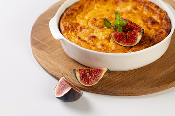 curd casserole with pumpkin in a round white shape on a wooden board for serving on a white table decorated with figs
