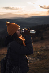 A young woman in an orange beanie hat is drinking tea from a thermos outdoors