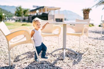 Little girl stands between a chair and a table on a pebble beach. High quality photo