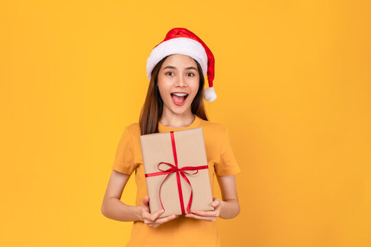 Cheerful beautiful Asian woman wearing christmas hat with stand with holding gift box with red ribbon on light yellow background.