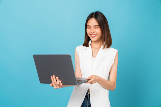 Studio shot of cheerful beautiful Asian woman holding laptop with typing on keyboard on blue background.